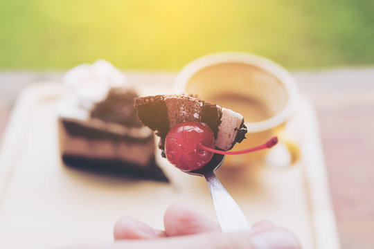 Warm Tone Photo Of Hand Holding Spoon Of Cherry And Chocolate With Cake And Coffee Cup Background