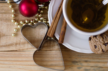 Cup of green tea, two cinnamon sticks, chocolate cookies, two metal heart shaped  forms and christmas decoration on wooden background