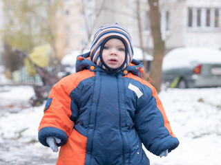 Attractive baby boy playing with the first snow. He smiles and looks snowman. Thick blue-orange jumpsuit bright striped hat on a year-old child.