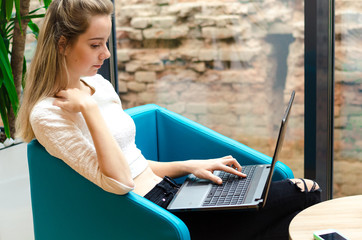 Portrait of beautiful smiling woman sitting on a comfortable chair in a cafe with black laptop. Pretty student doing work with laptop.