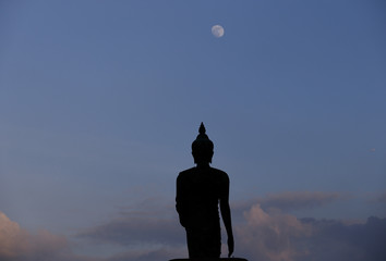 A moon shines in a sky over a large silhouetted Buddha statue in Bangkok, Thailand.