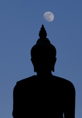 A moon shines in a sky over a large silhouetted Buddha statue in Bangkok, Thailand.