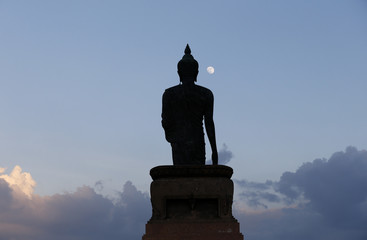 A moon shines in a sky over a large silhouetted Buddha statue in Bangkok, Thailand.