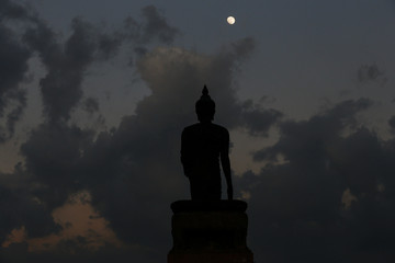 A moon shines in a sky over a large silhouetted Buddha statue in Bangkok, Thailand.