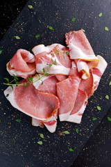 Raw smoked bacon slices on black stone board which herbs