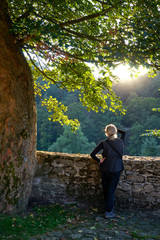 Fototapeta premium Man shading his eyes with his hands against the blazing sun under an old tree on a stone terrace with a low stone wall high above the wooded valley