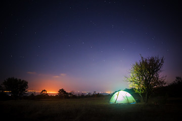 Camp tent stands on glade under starry night sky and shines from