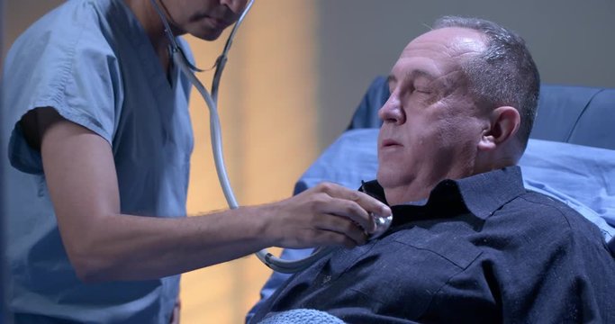 Male doctor uses stethoscope to check on mature male patient in hospital ward in the evening.  Medium close up recorded at 60fps