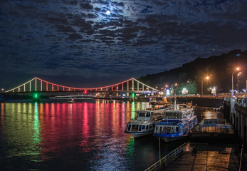 Fototapeta premium Glowing multicoloured city bridge above river and boats at quay