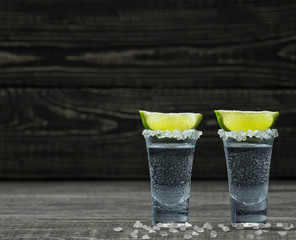 Two glasses of cold silver tequila in lime on a black wooden bac