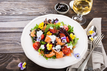 Fresh salad with smoked salmon, black olives, cherry tomatoes and edible flowers on wooden background.
