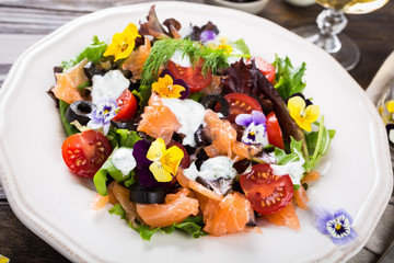 Fresh salad with smoked salmon, black olives, cherry tomatoes and edible flowers on wooden background.
