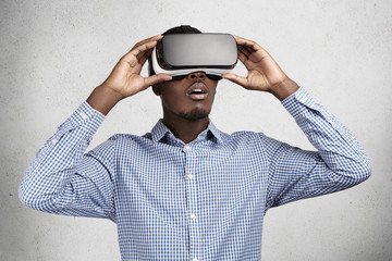 People, technology, innovation and gaming concept. Astonished African businessman in checkered shirt experiencing virtual reality while using oculus rift headset for entertaining himself in office