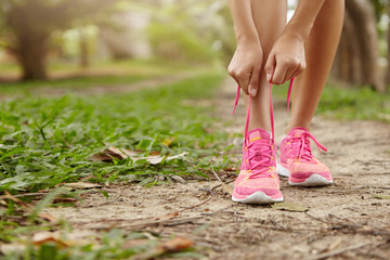 Caucasian athletic woman tying laces on her pink running shoes before jogging standing on footpath in forest. Female runner lacing her sneakers while doing workout in rural area. Film effect