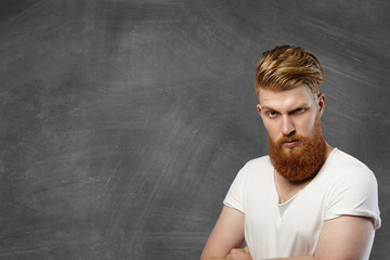 Headshot of hipster fashionable man with long red beard and stylish haircut having angry and unhappy facial expression, scowling and frowning while posing against blank blackboard with arms crossed