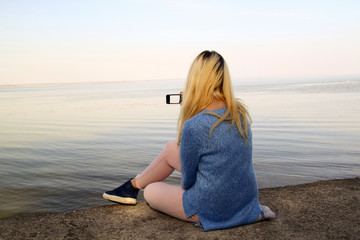 Fototapeta na wymiar Young girl photographed on a mobile phone sitting on the shore o