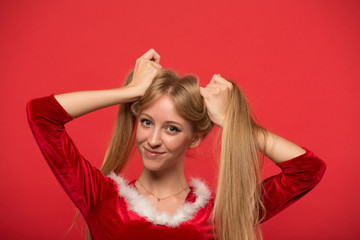 Naklejka premium Beautiful young woman in Santa costume playing with a strand of her long silky hair, looking at camera on red background