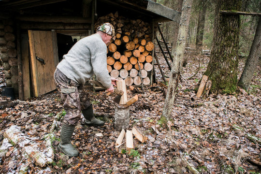 Man Chopping Wood. Cloudy, Snow On The Ground. Wooden House In The Forest. Hunting Lodge. A Man Of 50 Years. A Man Dressed In A Sweater And Hat.