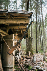 Hunting lodge in the forest. At the entrance hangs a hunting rifle. Cloudy weather on earth is some snow. Near the wooden house stacked firewoods.