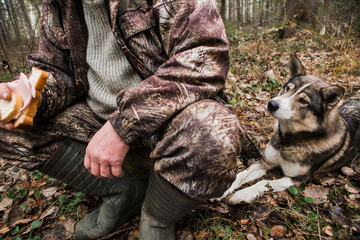 Male hunter in the forest eat a sandwich. Next to the man rests his hunting dog and looks at the sandwich. Hunters at Rest. Cloudy, autumn. The man with the dog.