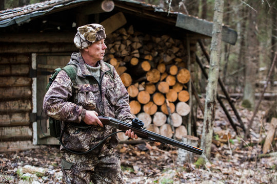Man the hunter is in the forest in rubber boots, with a backpack and a gun. Cloudy weather, autumn. Against the background is seen the wooden lodge. Around the house are wood for the fire.