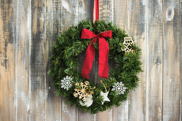Fototapeta na wymiar Christmas aged wood background. Traditional pine xmas decoration at brown wooden door or wall. Pine Christmas wreath decorated with toys, red ribbons hanging in home interior. Horizontal colour photo.
