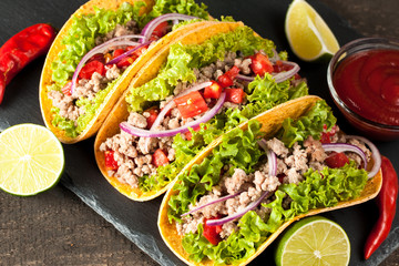 Photo of Mexican tacos with ground meat, beef, beans, onions and salsa on wooden background. Ketchup sauce and lime. A glass o beer in the background.