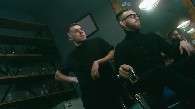 4K CINEMAGRAPH - Young satisfied Caucasian man sitting with whiskey after a getting haircut in a modern barber shop. Seamless loop. 4K UHD Raw edited footage