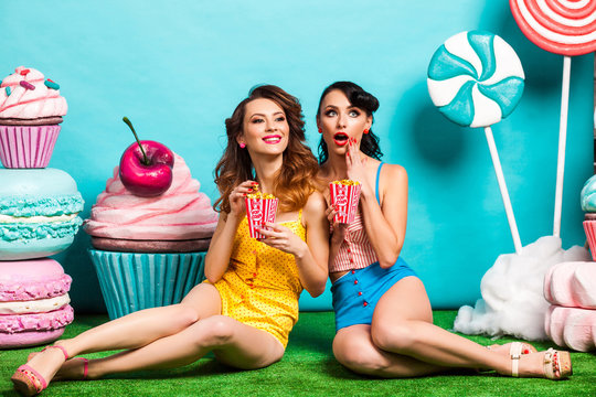 Two young cool lady on turquoise background, sitting on the grass and eating popcorn, laughing, best friends, crazy emotions, Huge candy, lollipops, cotton candy, Cake, fashionable Pin-up girl