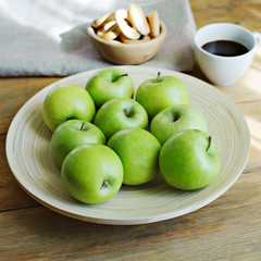 Green apples, donut on a napkin and coffee on wooden background, square.