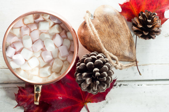 Hot chocolate, topped with marshmallow and served in a shiny copper mug. On a white wooden table with autumn leaves, pine cone and wooden table decoration.