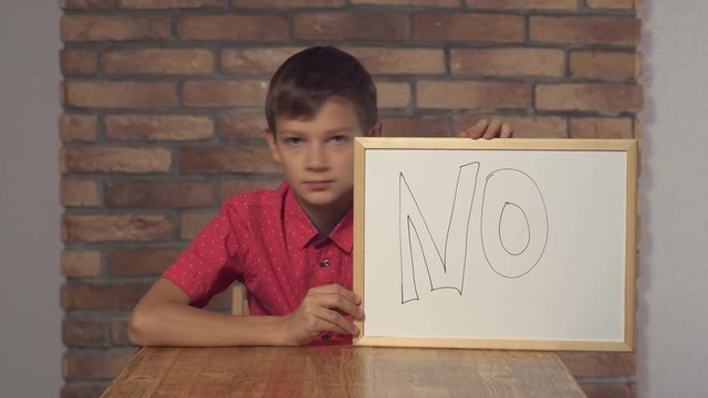 Child sitting at the desk holding flipchart with lettering nop on the background red brick wall.