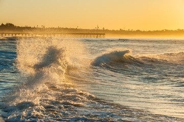 Back washed waves collide and smash with Ventura Pier in glowing background. 