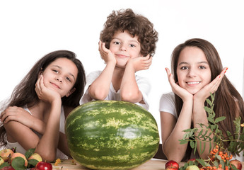 Two girls and a boy with his head on his hands, sitting at the table in front of a large watermelon whole. Looking at the camera. White background. Close-up. © kravik93
