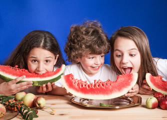 Little curly boy and two girls sitting at a wooden table and eat large ripe watermelon. Blue background. Close-up.