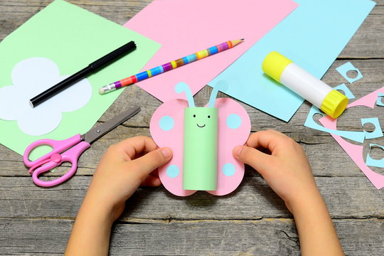 Child holding colored paper butterfly in hands. Child shows a fun paper diy. Stationery on an old wooden background. Preschool paper and glue crafts activities. Children creative abilities development