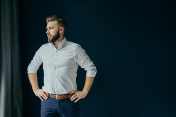 View in profile of a young bearded businessman, dressed in a white shirt standing near the window on a dark-blue walls and looking out the window. - obrazy, fototapety, plakaty