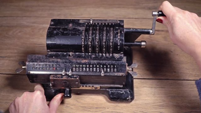 Female hand turning the handle of the old, rusty adding machine, standing on a wooden table