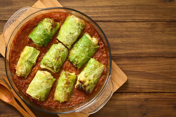 Vegetarian stuffed savoy cabbage rolls filled with wholegrain rice, pepper, onion and carrot, baked on tomato sauce in pan, photographed overhead on dark wood with natural light