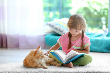 Red cat and cute little girl reading book on carpet - Powered by Adobe