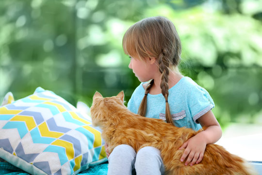 Cute Little Girl With Red Cat Sitting On Bed Beside Window