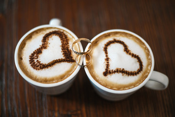 wedding rings and two cups of coffee with foam in the form of heart. Wooden background.