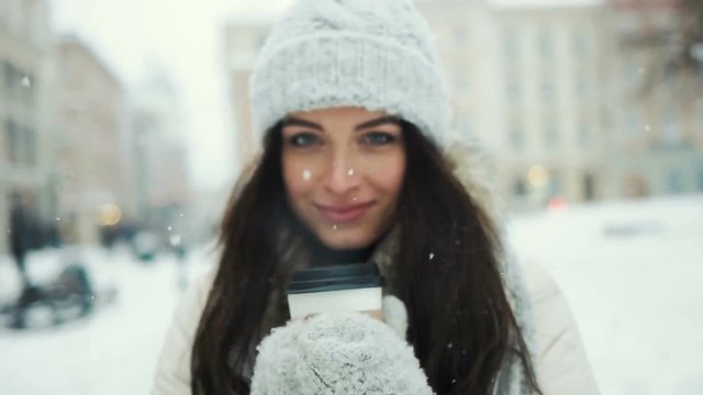 happiness, winter holidays, christmas, beverages and people concept - smiling young woman in white warm clothes with and drinking coffee to take away over snowy city background