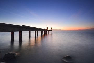 Fototapeta na wymiar Beautiful dusk sky over the long jetty with silhouette of anglers. Long exposure shot of seascape.