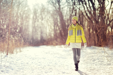 Winter happy woman walking in snow outdoors nature. Joyful young person relaxing on an outdoor walk activity in snowy forest landscape wearing warm yellow fashion outerwear jacket boots, scarf, hat.