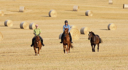 Two Women Horseback Riding in a Field with Bales of Hay