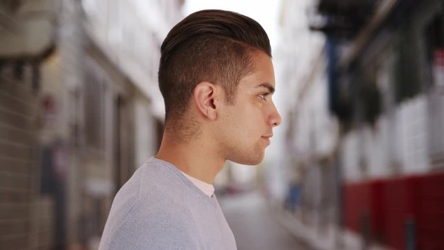 Side view of Hispanic man with cool undercut standing in alley in city. Profile close up portrait of Latino millennial