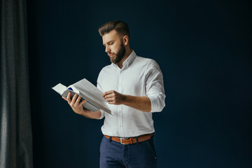 View in profile of bearded businessman,dressed in white shirt standing near window on dark blue wall and reading book.Man stands and leafing through thick book,which is in his hands.Man holding book.
