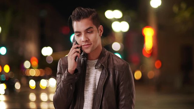 Fashionable millennial Latino man talking on smartphone on city street at night. Hispanic guy using cell phone to make call smiling