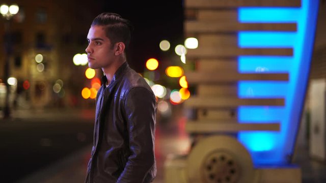 Fashionable millennial Latino man standing on city street at night. Portrait of handsome Hispanic guy wearing leather jacket in the evening